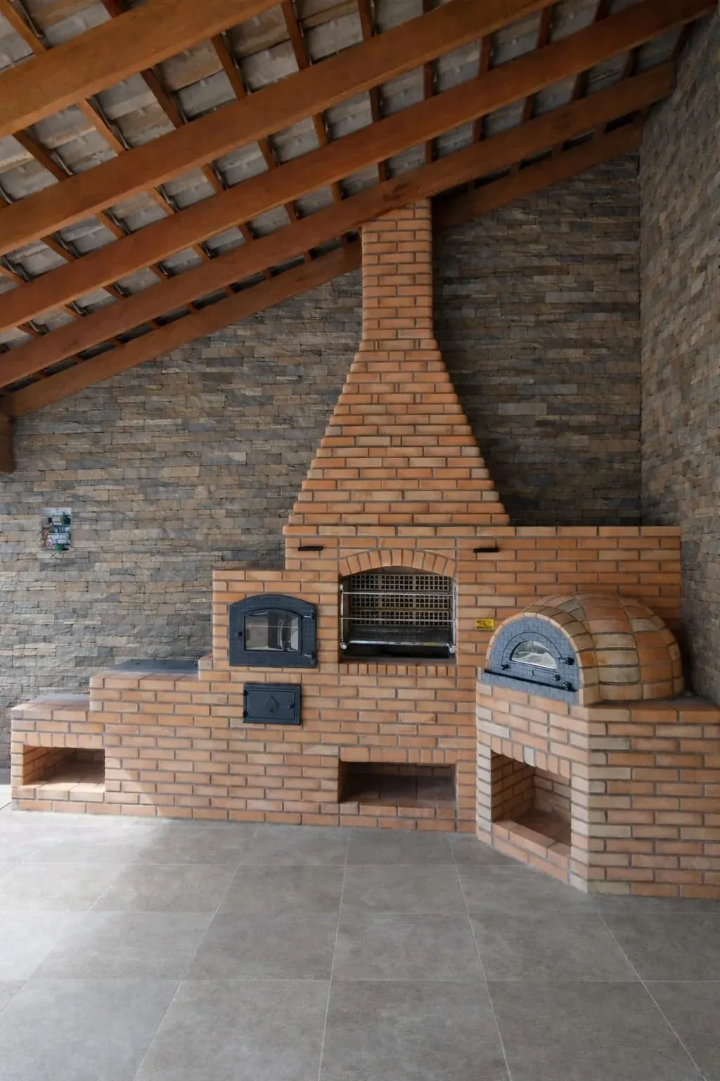 Conjunto 4 em 1 de tijolos com churrasqueira, fogão a lenha, forno de ferro fundido e forno igloo (pizza) sob cobertura de madeira.