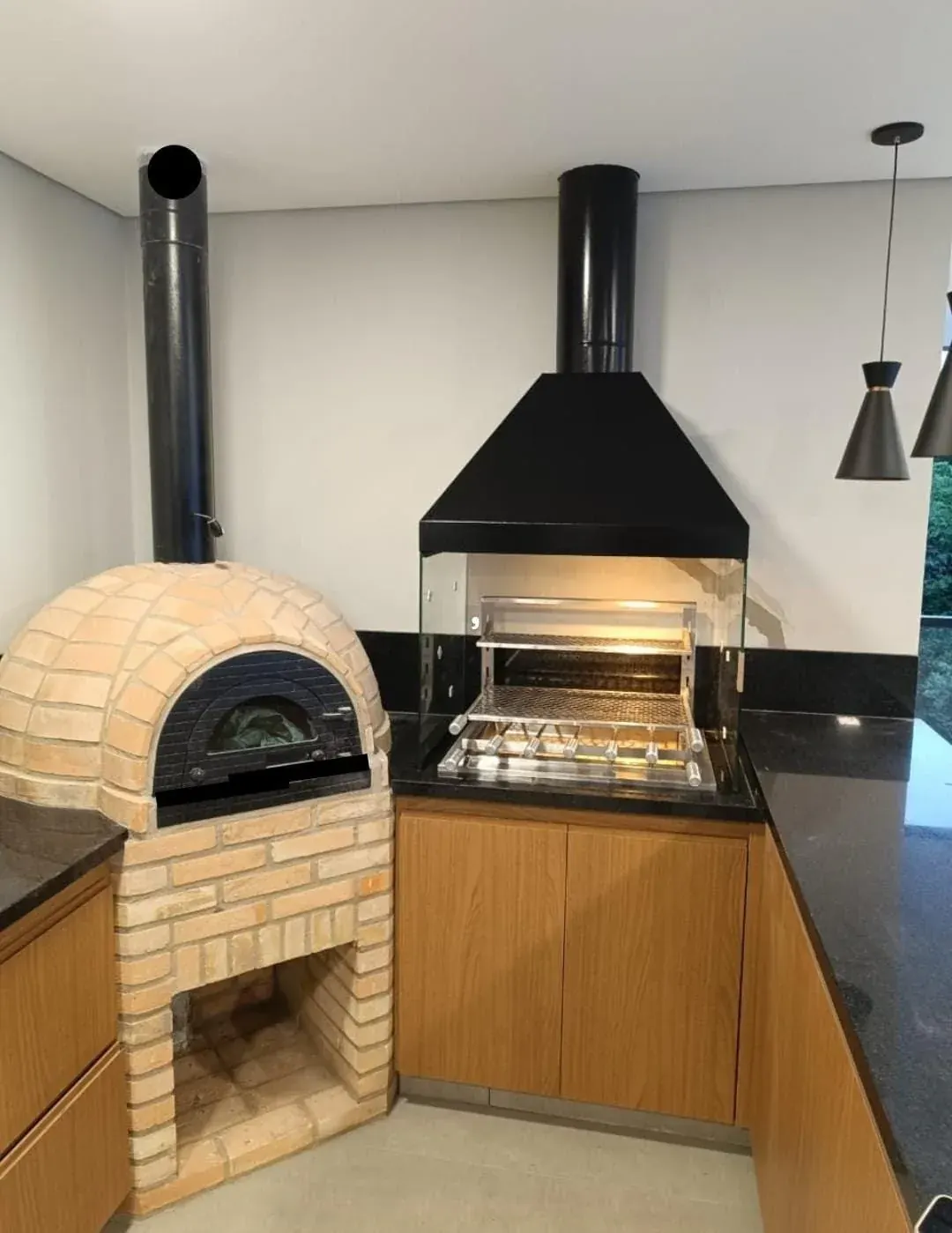 Forno igloo (pizza) ao lado de churrasqueira com coifa escura e painéis de vidro.